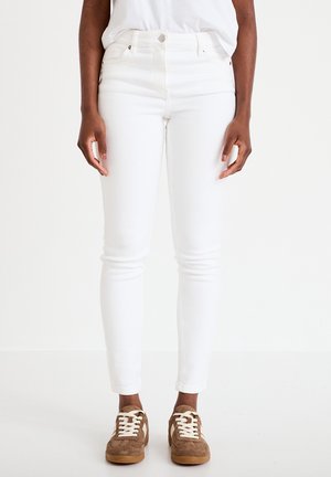 Person iført hvide slim-fit jeans og brune ruskinds sneakers, stående mod en ensfarvet hvid baggrund.