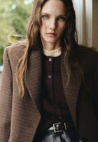 Blazer de tweed marrón con un diseño estructurado, superpuesto a un suéter de punto granate con botones, y un cinturón negro que acentúa la cintura.