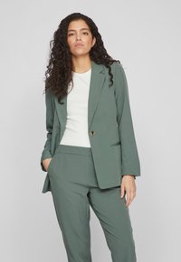 Tmavě zelený elegantní oblek skládající se z blazeru a odpovídajících kalhot, vyrobený z hladké tkaniny s jedním knoflíkem a klopovými kapsami.
