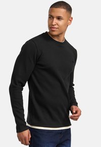 Camisa negra de manga larga con textura acanalada, cuello redondo y un pequeño logo en el pecho. Llevada sobre una capa blanca y combinada con pantalones vaqueros azules.