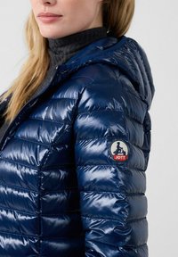 Giacca imbottita blu navy con finitura lucida, dotata di cappuccio e una toppa circolare con logo sulla manica. Design a righe orizzontali.