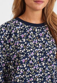 Frau in einem marineblauen Oberteil mit einem mehrfarbigen abstrakten Blumenmuster in Weiß, Pink, Blau und Grün, mit einem marineblauen Halsausschnitt und langen Haaren.