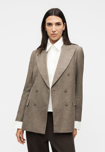 Γυναίκα με μακριά σκούρα μαλλιά που φοράει μπεζ διπλόμπουλο σακάκι πάνω από λευκό πουκάμισο με γιακά και σκούρο παντελόνι, στέκεται μπροστά σε ένα απλό φόντο.