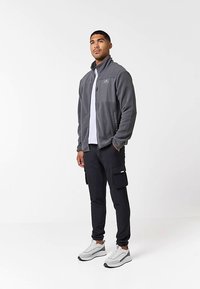 Uomo in piedi, con una giacca in pile grigia con zip, una maglietta bianca, pantaloni cargo neri e scarpe da ginnastica bianche, su uno sfondo bianco semplice.