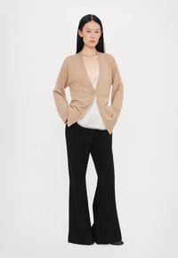 Beige kötött kardigán geometrikus mintával, nyitott elülső résszel és egy gombos részlettel, fehér top és fekete bőszárú nadrág felett viselve.