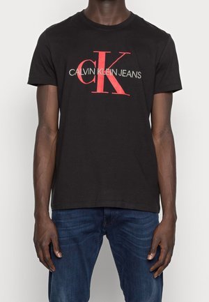 Mężczyzna ubrany w czarny t-shirt Calvin Klein Jeans z czerwonym logo "cK" i szarym napisem, w zestawieniu z niebieskimi dżinsami, stojący na jednolitym tle.