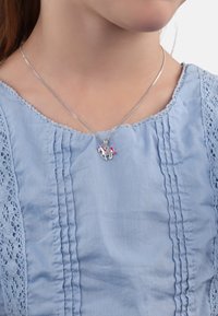 Srebrny naszyjnik z zawieszką w kształcie jednorożca, z różową grzywą, noszony na jasnoniebieskiej, fakturowanej bluzce z koronkowymi akcentami.