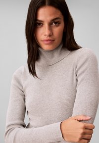 Femme aux cheveux raides et foncés portant un pull à col roulé côtelé gris clair, regardant la caméra les bras doucement croisés.