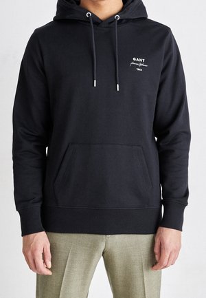 Mann trägt einen schwarzen GANT Hoodie mit Fronttasche und Kordeln, kombiniert mit hellen Hosen, steht vor einem einfarbigen Hintergrund.