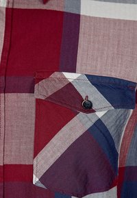 Närbild av en rutig skjortficka i röd, blå och vit färg med en klaff som är säkrad med en svart knapp.