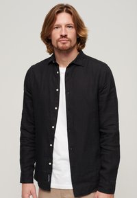 Camisa larga negra de manga larga hecha de una tela ligera con un cierre de botones al frente, botones blancos y una textura sutil. Llevada sobre una camiseta blanca.