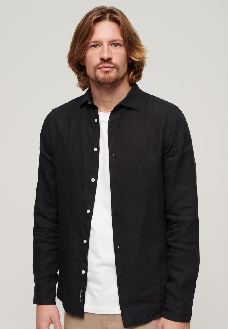Camisa larga negra de manga larga hecha de una tela ligera con un cierre de botones al frente, botones blancos y una textura sutil. Llevada sobre una camiseta blanca.