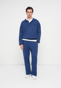 Μπλε φούτερ με γιακά και τέσσερις κουμπιά, σε συνδυασμό με ταιριαστές μπλε παντελόνες. Και τα δύο είναι φτιαγμένα από απαλό ύφασμα. Λευκά αθλητικά παπούτσια.