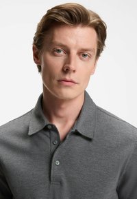 Junger Mann mit hellbraunem Haar, der ein graues Hemd mit Kragen trägt, vor einem schlichten weißen Hintergrund.