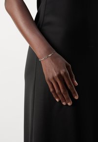 Silverarmband med länkdesign och en intrikat accent. Bärs på en hand mot en svart klänning.