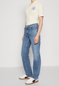 Camisa polo beige con logotipo azul, combinada con jeans rectos de mezclilla azul claro y zapatillas blancas con acentos negros.