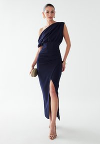 Vestido azul marino de un solo hombro con cuerpo drapeado, cintura ajustada y abertura lateral hasta el muslo; hecho de material elástico; combinado con un bolso de mano y tacones.