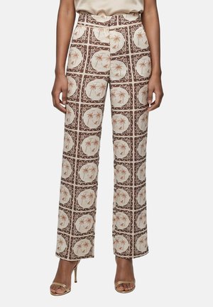 Pantalon femme taille haute à jambes larges avec motif de palmiers marron et beige, porté avec un haut beige et des sandales à talons hauts dorées.