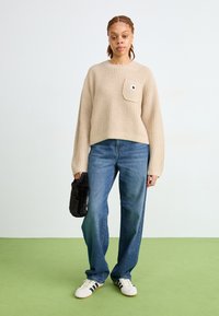 Maglione beige lavorato a maglia con tasca frontale, jeans in denim blu a gamba larga e scarpe bianche con strisce scure. Una borsa nera in ecopelliccia è tenuta in mano.