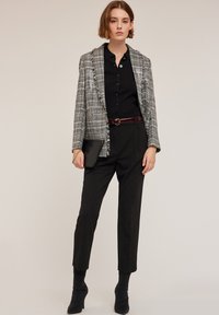 Grå rutig kavaj med fransdetaljer över en svart skjorta, tillsammans med svarta skräddarsydda byxor och klackade ankelstövlar, hållandes en liten clutch.