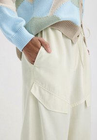 Lichtbeige broek met voorzakken, gecombineerd met een gebreide trui in blauw, groen en beige met een patroon. De stof lijkt zacht en gestructureerd te zijn.