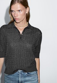 Ciemnoszara dziergana koszulka polo z fakturowanym wzorem, zapinanym kołnierzykiem i krótkimi rękawami, zestawiona z jasnoniebieskimi dżinsami.