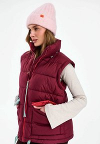 Nő rózsaszín kötött sapkában, bordó pufi mellényben világos bézs hosszú ujjú felső felett, kezét a mellény zsebében tartva, oldalra néz.