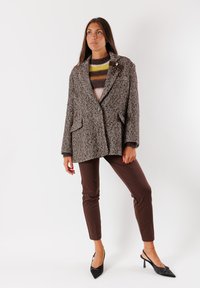Blazer oversize marrone con motivo a scacchi, dotato di due grandi tasche anteriori, chiusura con bottone e dettaglio decorativo a fiocco. Abbinato a pantaloni marroni.