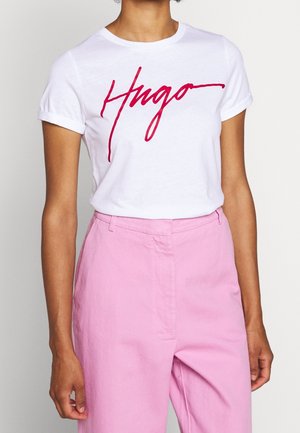 Personne portant un t-shirt blanc avec le texte rouge "Hugo" et un pantalon rose clair taille haute, debout devant un fond uni.