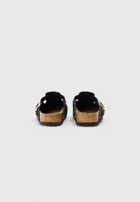 Sandales noires en suède sans dos avec semelle en liège, accents en métal doré et semelle texturée. Design à talon ouvert avec deux brides réglables.