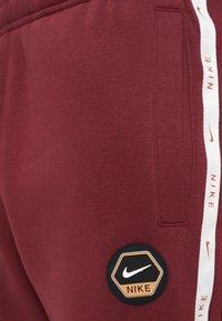 Burgundijas krāsas sporta bikses ar strukturētu virsmu, sānu kabatām un sešstūra Nike logotipa uzšuvi ar apelsīna akcents. Baltā krāsa apzīmējumā gar sānu.