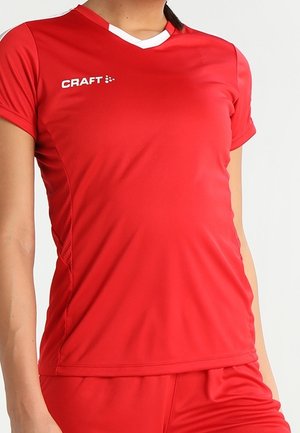Kvinde iført en rød sportstrøje med korte ærmer og matchende shorts med hvid kantet V-halsudskæring og "CRAFT"-logo på brystet.