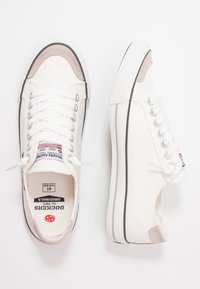 Weisse Canvas-Sneaker im Low-Top-Stil mit beigen Akzenten. Merkmale sind eine runde Zehenpartie, weisse Schnürsenkel und Gummisohlen mit einem kontrastierenden schwarzen Rand.