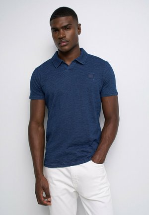 Jeune homme portant un polo bleu à manches courtes et un pantalon blanc, debout devant un fond blanc uni avec une main dans la poche.