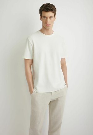 Młody mężczyzna z krótkimi kręconymi włosami, ubrany w białą, teksturowaną koszulę z krótkim rękawem i jasnobeżowe spodnie, stojący z rękami w kieszeniach.