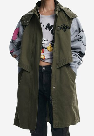 Personne portant un manteau sans manches vert par-dessus un sweat-shirt gris Mickey Mouse, associé à un jean noir taille haute, les mains dans les poches.