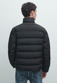 Black puffer jacket with horizontal quilted patterns, high collar, and elastic cuffs. Worn over denim jeans, viewed from the back.