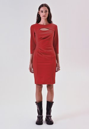 Rotes, figurbetontes Kleid mit Three-Quarter-Ärmeln, das einen vorderen Ausschnitt, geraffte Textur und einen knielangen Saum hat. Getragen mit schwarzen kniehohen Stiefeln.