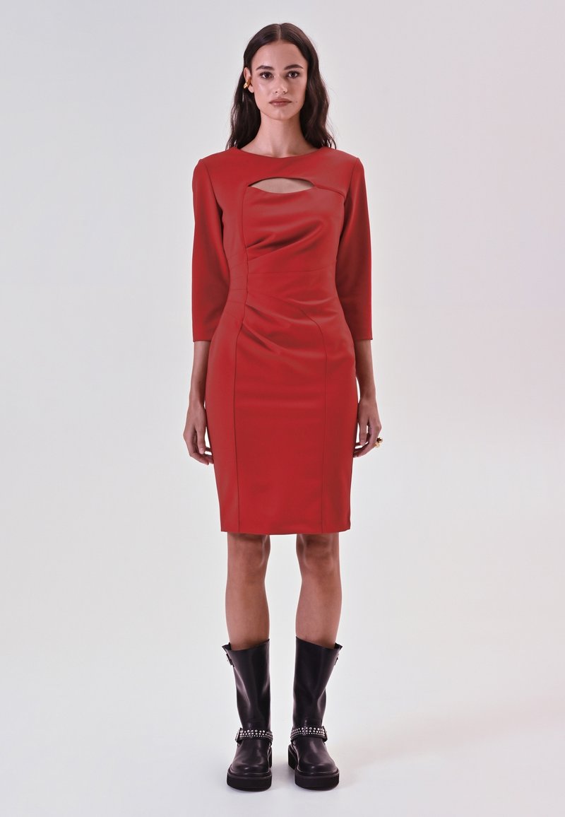 Rotes, figurbetontes Kleid mit Three-Quarter-Ärmeln, das einen vorderen Ausschnitt, geraffte Textur und einen knielangen Saum hat. Getragen mit schwarzen kniehohen Stiefeln.