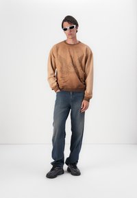 Bruine sweatshirt met een adelaar graphic, lichte naar donkere gradient. Gecombineerd met blauwe jeans en zwarte sneakers. Accessoires met witte zonnebril.
