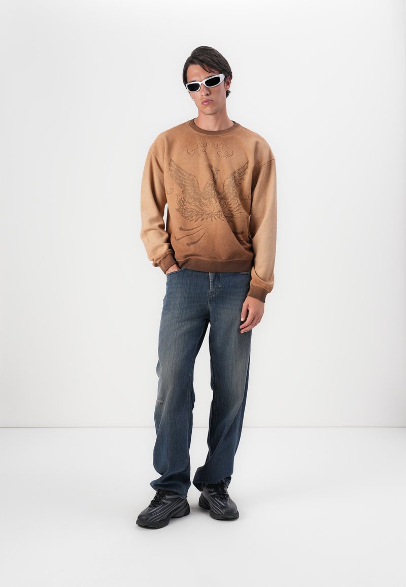 Bruine sweatshirt met een adelaar graphic, lichte naar donkere gradient. Gecombineerd met blauwe jeans en zwarte sneakers. Accessoires met witte zonnebril.