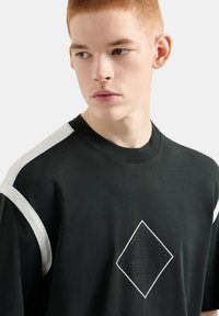 T-shirt nero a maniche corte con accenti bianchi sulle spalle e un'etichetta a forma di diamante testurizzata con lettere in rilievo al centro.