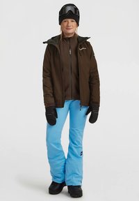 Donna che indossa una giacca marrone, pantaloni da neve blu, guanti neri, un berretto nero e occhiali da sci, in piedi con un leggero sorriso su uno sfondo semplice.