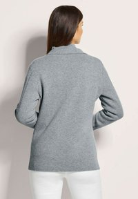 Grijze gebreide trui met een hoge kraag, lange mouwen en een relaxte pasvorm. De stof lijkt zacht en textuur te hebben, met zijsplitten aan de zoom.