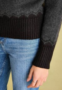 Genähter Pullover in Farbverlaufsschattierungen von Grau und Schwarz, mit gerippten Ärmelbündchen und einem gewellten Saum. Kombiniert mit hellblauen Jeans.