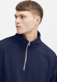 Pull en polaire marine avec un col haut, fermeture éclair, et un petit logo en relief sur la poitrine. Texture lisse et coupe décontractée.