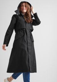 Long black raincoat with a hood, drawstring waist, and flap pockets. Made from waterproof material with a smooth texture.
