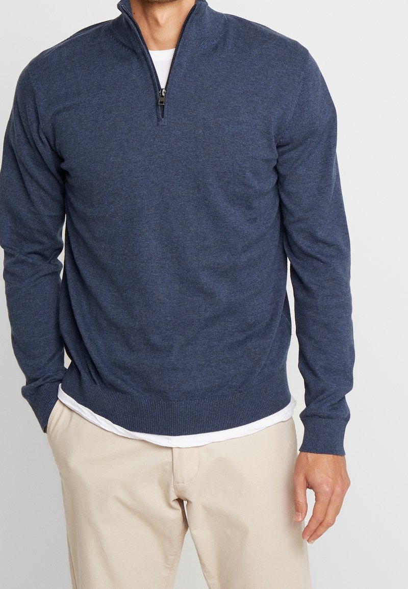 Homme portant un pull bleu marine à fermeture éclair au quart sur une chemise blanche, associé à un pantalon beige, debout avec une main dans la poche.