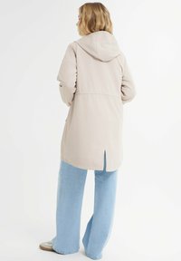 Giacca beige con cappuccio, vita cintrata, maniche lunghe, tasche laterali e uno spacco all'orlo, abbinata a jeans a gamba larga blu chiaro.