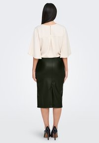 Blouse beige avec des manches courtes et larges et un pli au dos. Jupe crayon vert foncé en tissu brillant. Chaussures à talons noirs.